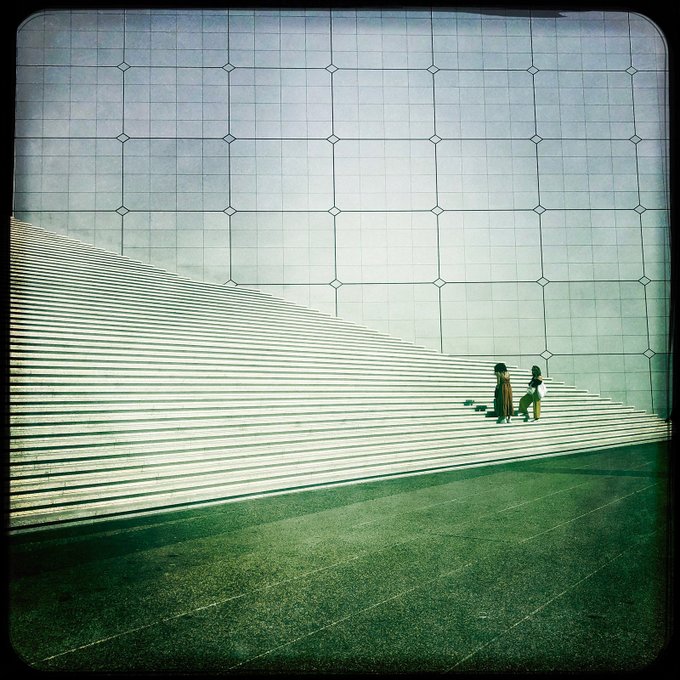 The image features a couple walking up a set of stairs with white lines marking their path. The stairs are part of an architectural structure, possibly a building or a bridge, which adds depth to the scene. There are two people visible on the steps - one person is closer to the left side and another person is located more towards the right side. They appear to be walking together in harmony as they ascend the stairs. The image also contains text that reads "GM Happy Saturday Le Parvis de la Défense A quiet urban geometry of scale, distance, and light." This text provides context about the location or time when this photo was taken, but it does not provide any additional information about the couple walking up the stairs.
