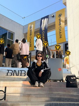 A person in sunglasses sitting in front of a building displaying 'BNB CHAIN' banners on yellow flags.