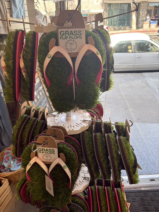 A retail display rack features 'GRASS FLIP FLOPS' with tags reading 'XS,' stocked with flip-flops that have green grass-like tops and various colored straps, displayed near a store window.
