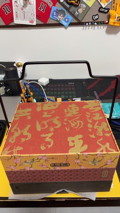 A decorative red and gold Asian-style gift box sits on a desk next to a computer setup and keyboard, with various sports-themed items pinned to the wall behind it.