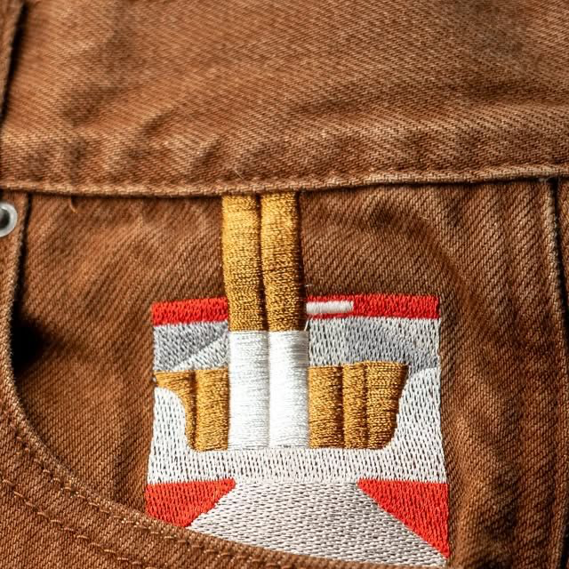 A close-up of an embroidered pocket on brown denim fabric, featuring a stylized design in red, white, gray, and gold/yellow thread with no visible text or numbers.