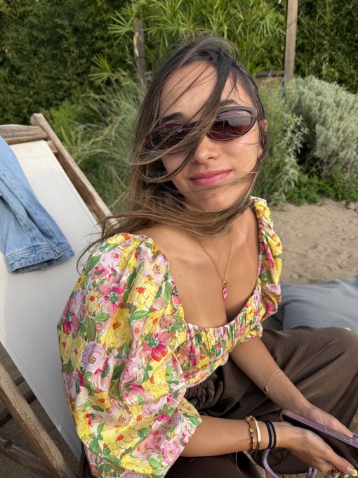 A woman wearing a colorful floral puff-sleeve top and oval sunglasses sits on a beach chair holding a phone, with greenery in the background.