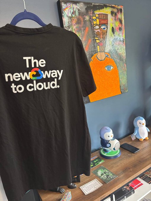 A black t-shirt with "The new way to cloud" text and a Google Cloud logo hangs next to a colorful abstract painting of an African figure, with penguin figurines on a wooden desk below.
