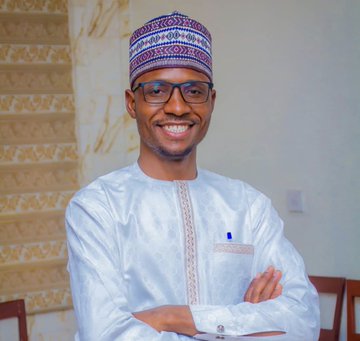 A smiling man wearing glasses, a patterned cap, and traditional white attire stands with his arms crossed in a brightly lit room. The image appears to be a portrait of Sunusi Minjibir, whose story of returning $135,000 inspired a viral meme coin in Crypto Twitter.
