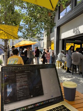 A laptop sits on a wooden table at an outdoor café under yellow umbrellas, with people gathered in the background.