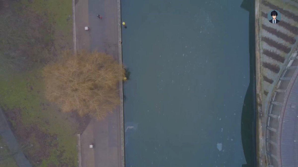 The image shows an aerial view of a city with a river running through it. The perspective is from above, providing a bird's eye view of the scene below. In the foreground, there are two people walking along the riverbank near a tree. The sky in the background appears hazy and gray, suggesting that the photo was taken during an overcast day or possibly on a cloudy day.