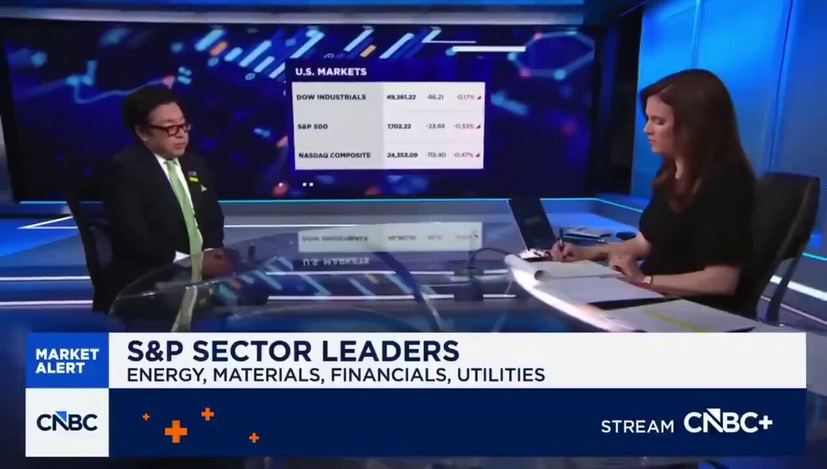 The image shows a man and woman sitting at a table, with a news segment playing on a television screen behind them. The news segment features an economic forecast for the next 18 months, which includes information about energy, materials, financials, utilities, and other industries. The man is wearing glasses, while the woman has long hair. They appear to be engaged in conversation or discussing the forecasts presented on the TV screen.