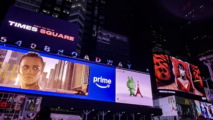 A nighttime view of Times Square's iconic illuminated billboards, including advertisements for Amazon Prime (featuring a cartoon character and Eiffel Tower) and Ernst & Young, with the 'TIMES SQUARE' sign and a 'Broadway' marquee visible.