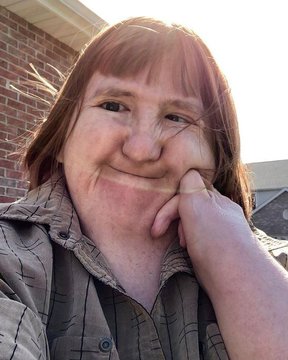 A close-up selfie of a smiling person with short reddish hair resting their chin on their hand, with a brick building in the background.