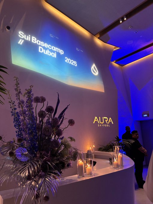 An indoor event space featuring a projection that reads 'Sui Basecamp // Dubai 2025' and a glowing 'AURA SKYPOOL' sign, with floral arrangements and candles in the foreground.