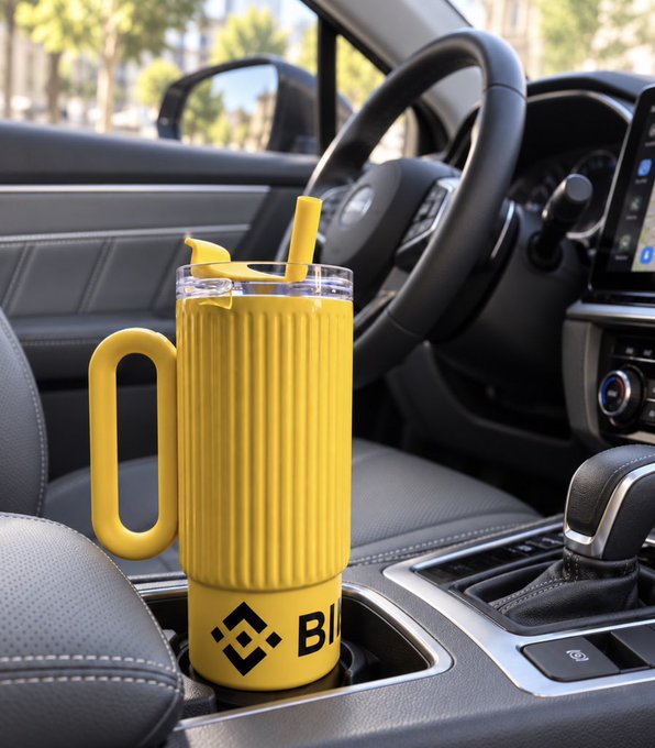 A yellow tumbler with a handle sits in the cup holder of a car's center console, featuring a Binance logo and partial text 'BI' on the bottom.