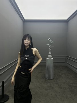 A young woman with long dark hair wearing a black cropped top and long black skirt standing in a minimalist gray room next to a decorative vase on a pedestal.