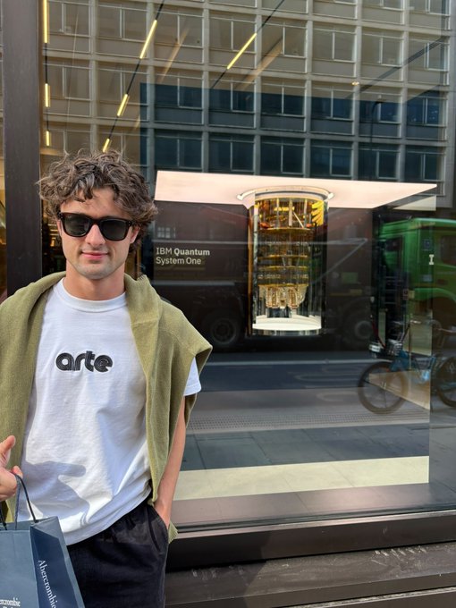 A man with curly hair and sunglasses wearing a white 'arte' t-shirt and green sweater stands in front of an IBM Quantum System One display, with a 'Alexander McQueen' shopping bag visible.