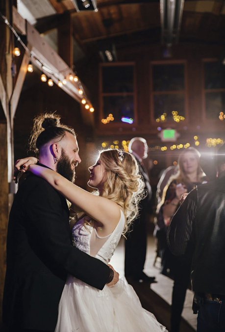 A man and woman in formal wedding attire dance closely together in a warmly lit venue decorated with string lights, while other guests watch in the background.