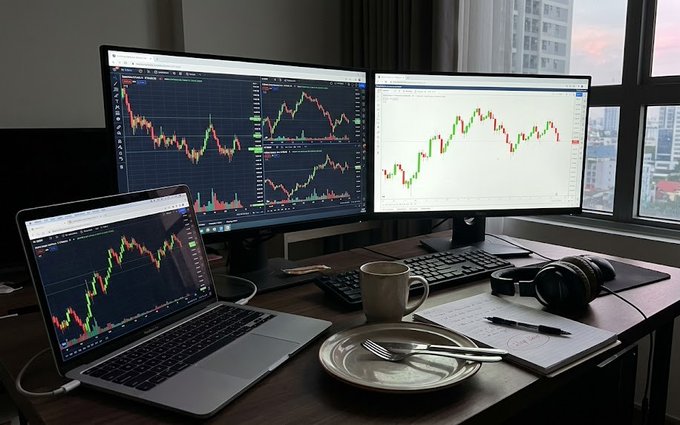 A trader's workspace featuring dual monitors and a laptop displaying candlestick charts, with a coffee mug, headphones, keyboard, and a plate with silverware on the wooden desk.