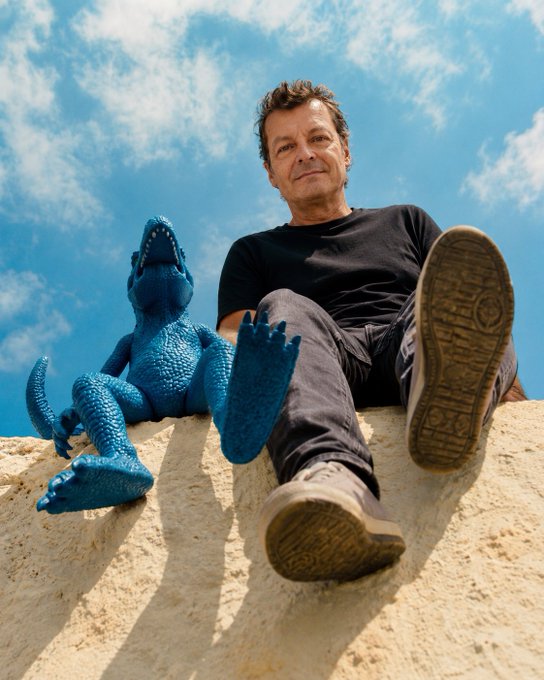 A man and a child wearing dinosaur slippers sit on a sandy dune against a bright blue sky, photographed from a low angle.