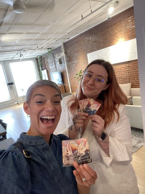 Two women take a selfie indoors, one holding up a trading card while wearing a denim top, the other in a white blazer with glasses.