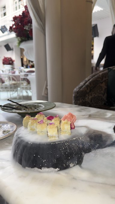 A plate of dessert cubes on a black stone base emitting white vapor, served on a marble table in what appears to be an upscale restaurant setting
