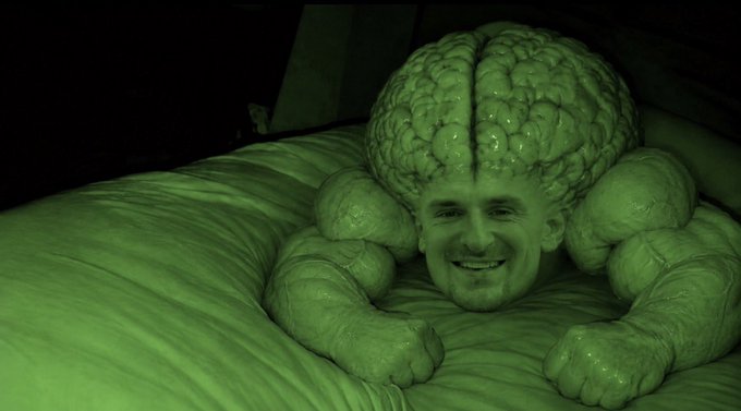 A person lies on a bed with a prop of an enormous brain enveloping their head and shoulders, illuminated in eerie green light.
