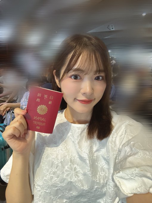 A young woman with long brown hair smiles at the camera while holding up a red Japanese passport ('JAPAN', 'PASSPORT') in what appears to be an airport setting.