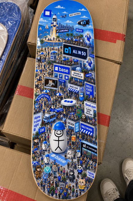 A colorful skateboard deck with detailed blue and white graphics featuring Base/GenLayer branding, crowds of characters, and phrases like 'ALL IN $0', 'BUILD EVERY DAY', and 'GENLAYER', resting on cardboard boxes.