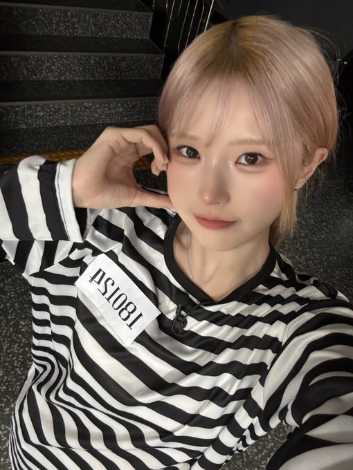 A selfie of a young woman with blonde/pinkish hair and bangs wearing a black and white striped shirt with a tag reading '1801SPA' visible, posing with her hand near her face.
