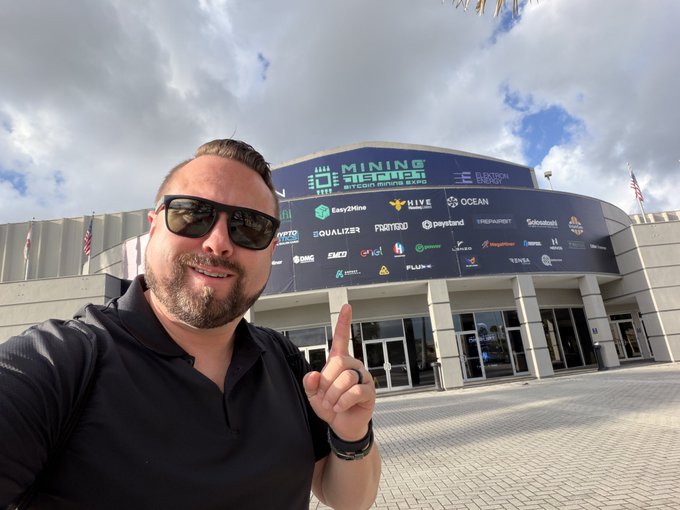 A man in sunglasses points excitedly at the Mining Disrupt 2024 banner above the venue entrance, which displays sponsor logos including Eterna Capital, Hive, Ocean, Easy2Mine, and Equalizer, capturing his arrival for Day 2 of the cryptocurrency mining conference.