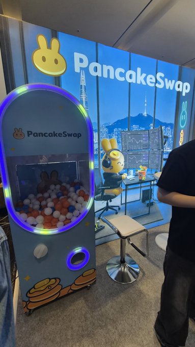 The image shows a PancakeSwap booth at Korea Blockchain Week, featuring a branded ball-prize machine with the PancakeSwap bunny logo, while the tagline "#KBWCAKE" and backdrop confirm the event setting mentioned in the post.