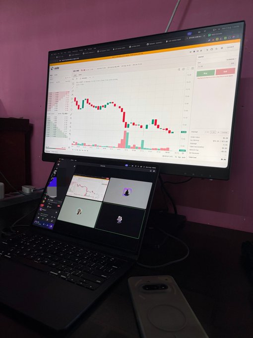 A desk setup showing a monitor displaying a stock trading chart with red and green candlesticks, with a laptop below showing a video call, plus a smartphone resting on the desk.