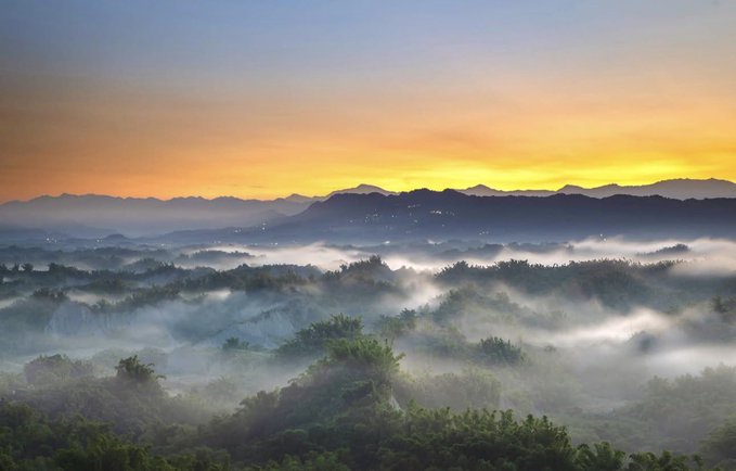 A misty sunrise or sunset over a forested landscape with rolling hills and mountains in the background under a golden sky.