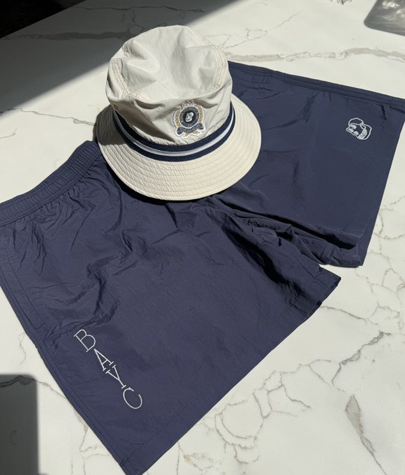 The photo displays navy blue shorts with a throwback look.

One pant leg reads the brand name "BATCH" vertically from bottom to top.  

A related hat with blue stripes sits above the shorts, designed for sunny days. The accompanying text mentions these "Bored Ape Yacht Club (BoredApeYC)" shorts, suggesting the owner is pleased with their timely arrival.