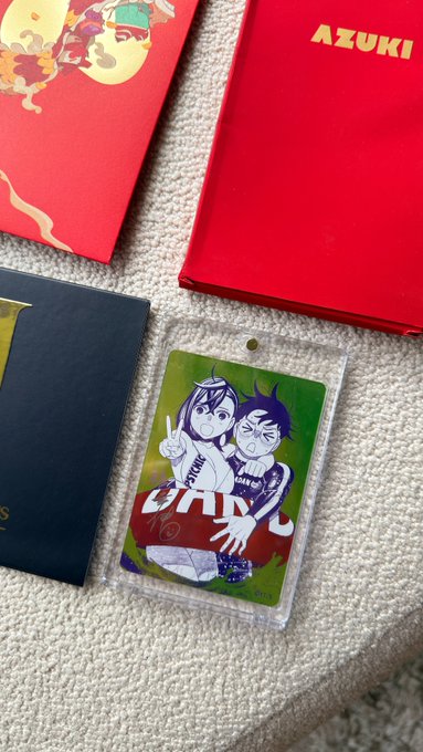 A photograph showing collectible card game merchandise on a white surface, including a red 'AZUKI' box and a green holographic card in a plastic sleeve featuring an illustration of two anime-style characters with 'DRM' text.