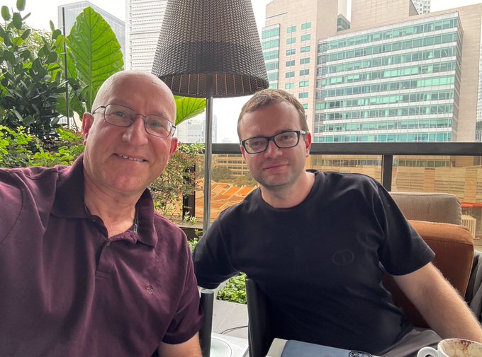 A selfie of two men wearing glasses sitting at a table on an outdoor patio with a city skyline of tall office buildings in the background.