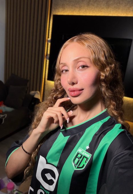 A selfie of a young woman with curly blonde hair wearing a green and black striped "B9 GAME" jersey.