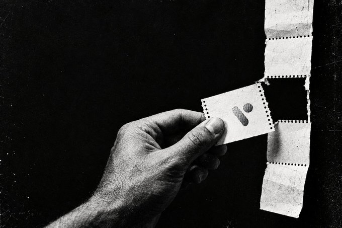 A black and white photograph showing a hand holding a perforated strip of paper with a symbol that resembles a lowercase 'i' or punctuation mark printed on it.