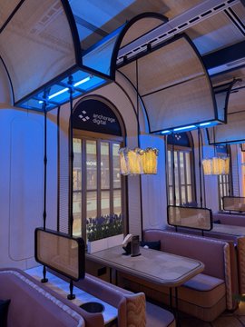 An interior view of an ornate dining car or lounge featuring blue lighting, arched windows, decorative chandeliers, and elegant table settings with cushioned booth seating.