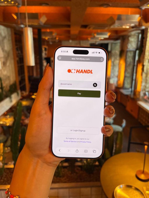 A hand holding a smartphone displaying the app.handlpay.com login page with 'Pay' and 'Login/Signup' buttons, taken inside a warmly lit restaurant.