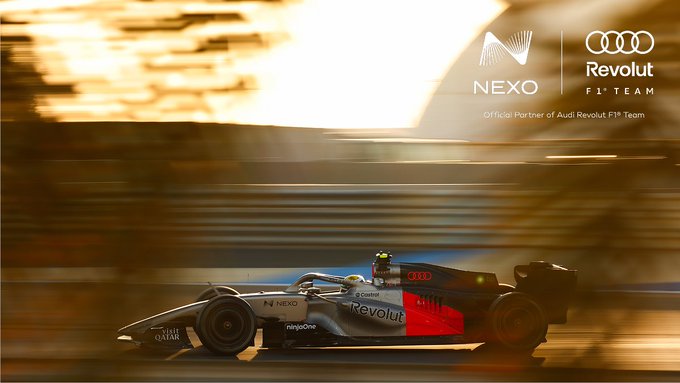 A Formula 1 car from the Audi Revolut F1 Team speeds along a track, bathed in the warm, golden light of the setting sun, with sponsorship branding for NEXO and "Official Partner of Audi Revolut F1 Team" displayed alongside. The motion-blurred background and low sun perfectly capture the "Golden hour. Full speed." caption, emphasizing the dynamic energy and dramatic lighting of the moment.