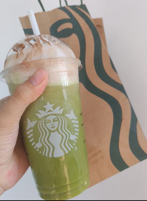 A hand holds a Starbucks cold cup filled with a green blended drink topped with foam and cocoa powder, in front of a brown Starbucks paper bag.