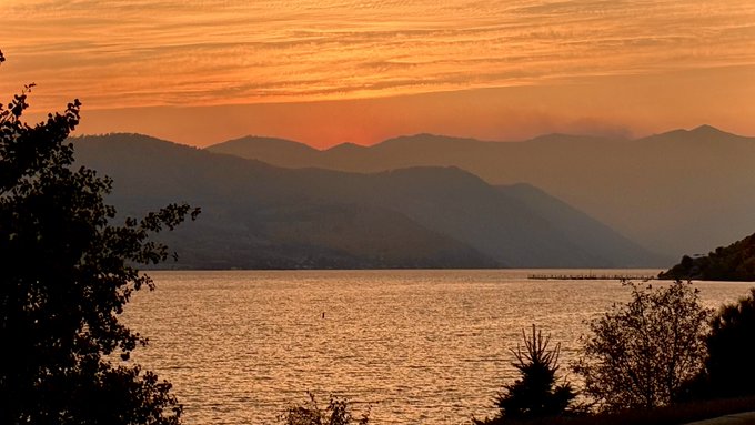 A serene sunset over a calm body of water with layered mountain silhouettes and silhouetted foliage framing the orange and pink sky.