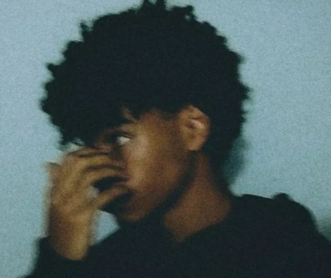 A grainy, low-light portrait shows a young person with curly hair, their hand raised near their face, wearing a dark shirt.