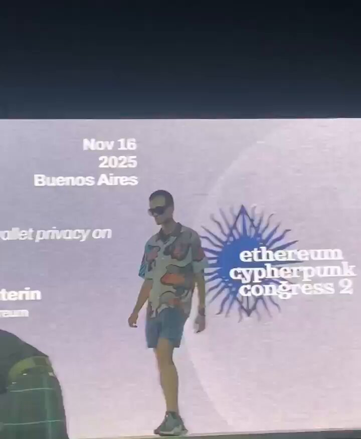 The image features a man walking down a runway with sunglasses on his head. He is wearing a shirt that has a unique design of clouds and rainbows, which adds a colorful touch to the scene. The background of the image shows a large screen displaying information about an event or gathering related to cryptocurrency. This suggests that the man might be participating in a fashion show or promotional event for a cryptocurrency-related project.
