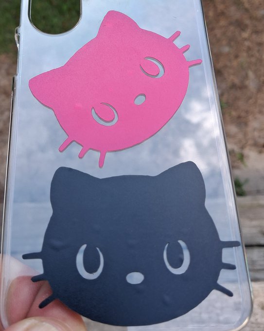 A clear phone case displays two cartoon cat face cutouts, one pink and one black, both featuring simple closed-eye expressions.