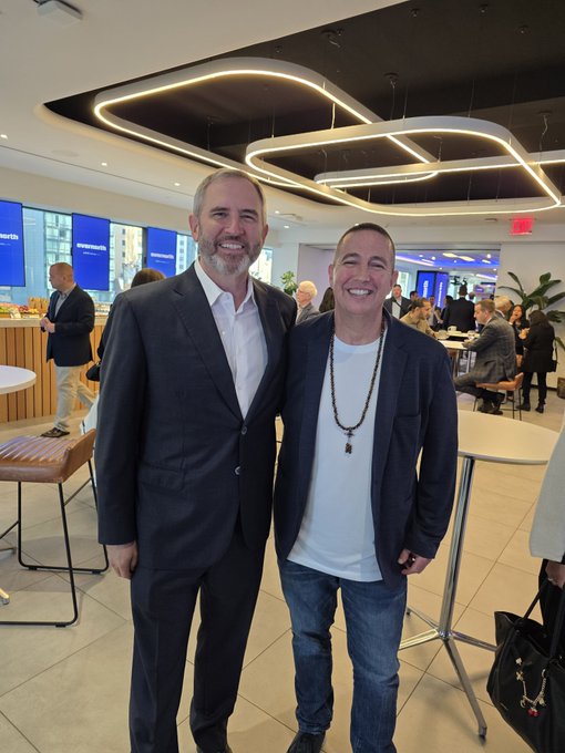 Two men smile and pose together in a modern lounge or event space with curved lighting fixtures and other people in the background.