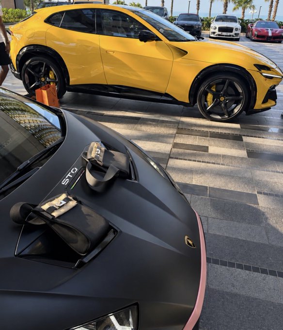 A matte black Lamborghini Huracán STO with designer bags on its hood is parked next to a yellow Ferrari Purosangue at a luxury car event, with "STO" branding visible on the sports car. The scene of exotic supercars symbolizes the aspirational wealth referenced in the post's bullish crypto prediction, suggesting that $SOL and $XRP gains will fund such luxury lifestyles.