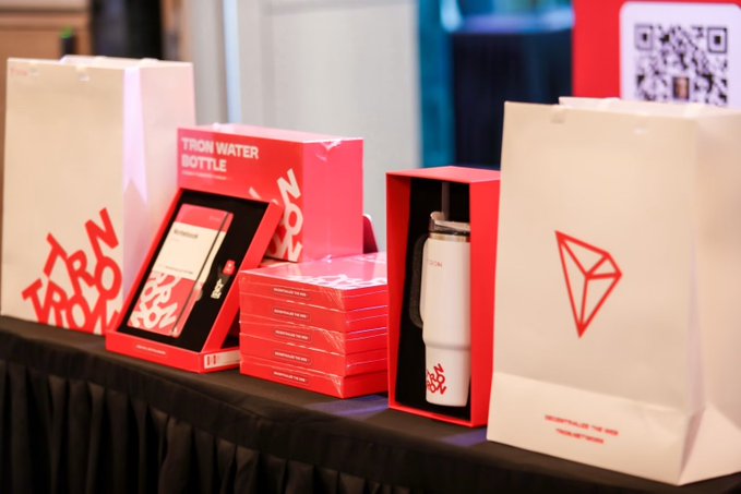 A table display featuring TRON-branded merchandise, including red and white "TRON WATER BOTTLE" boxes, a stainless steel bottle, and paper gift bags with the TRON logo, next to a QR code sign.