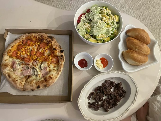 A spread of food on a round table including a pizza, salad, bread rolls, dipping sauces, and what appears to be grilled meat.