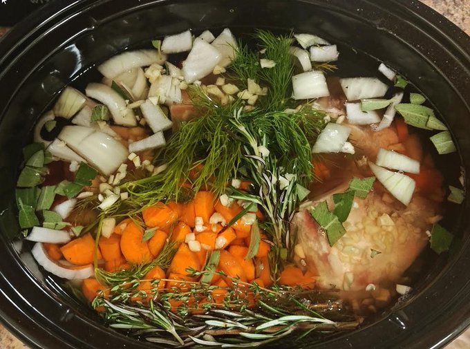 A slow cooker filled with ingredients for a meal, including chicken, chopped carrots, onions, garlic, and fresh herbs like rosemary, thyme, and dill in broth.