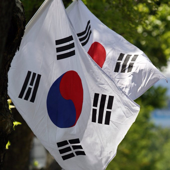 Two South Korean national flags (Taegukgi), with their iconic red and blue Taeguk symbol and black trigrams, flutter in the wind against a backdrop of green trees. The flags visually reinforce the post's headline about Jeonbuk Bank launching South Korea's first traditional bank Bitcoin-backed loans, symbolizing the country at the center of this financial milestone.