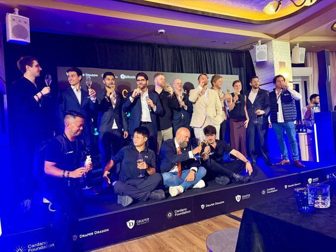 A group of around 15 people in business attire pose on stage with raised champagne flutes at a Draper Dragon and Mikobi event, with a backdrop featuring Cardano Foundation and Draper Dragon logos.